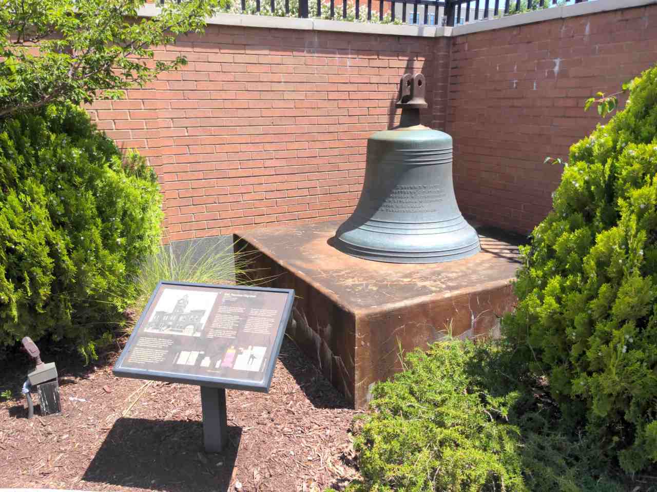 Large bell on block in background and historic marker in foreground.