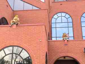 High above on brick building ledges are two Humpty Dumptys. One playing a guitar and the other using a spyglass.