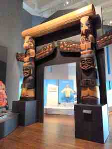 Portal between art exhibits is bordered and crowned by tall wood carved totem poles.