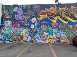 Building wall mural full of colorful graffiti and goofy looking cartoonish characters and creatures.